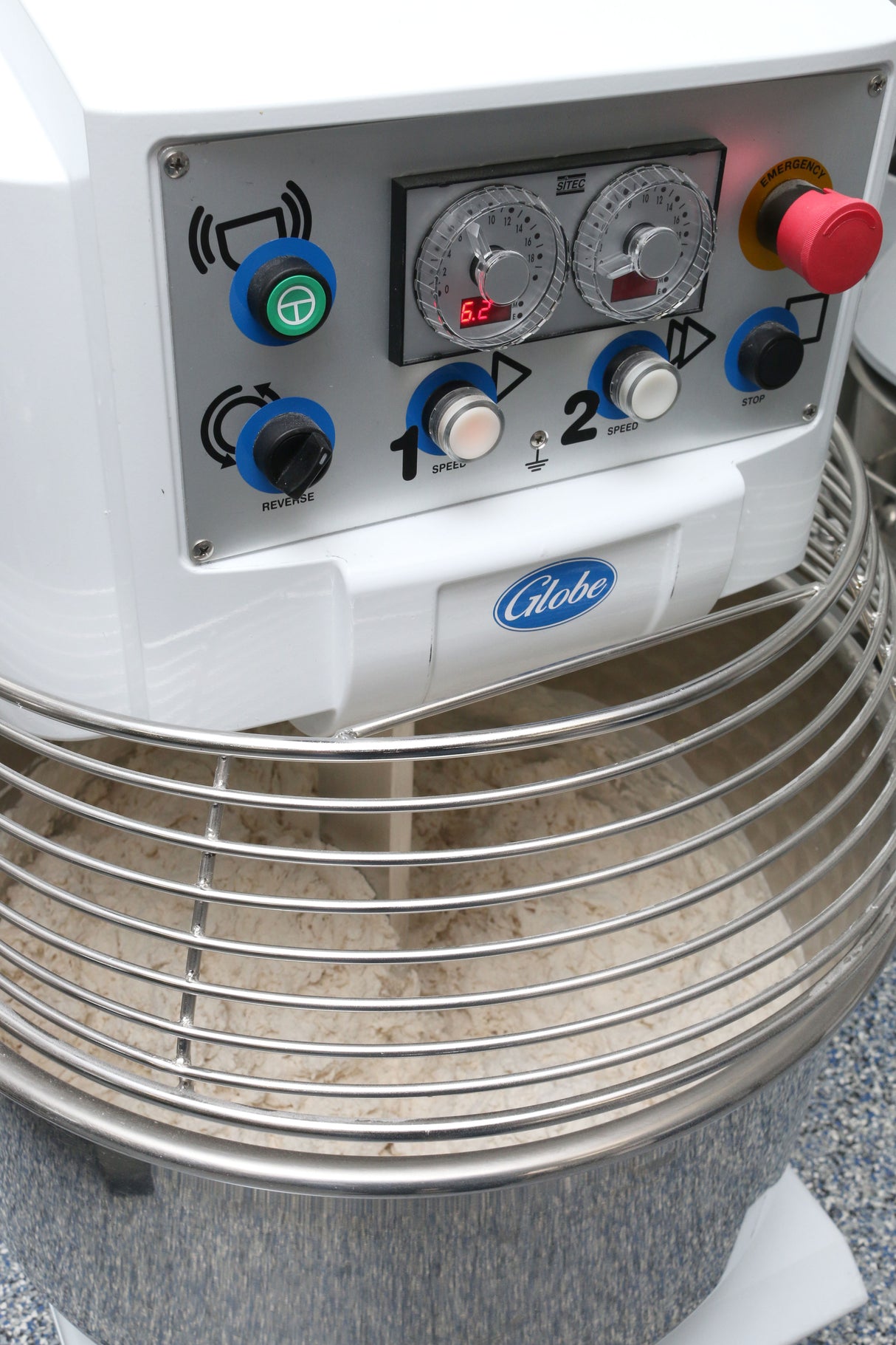 A close-up of a Globe spiral dough mixer with control knobs, buttons, and dials on the front panel. The stainless steel bowl below holds 130 lbs capacity of dough, protected by a wire safety guard.