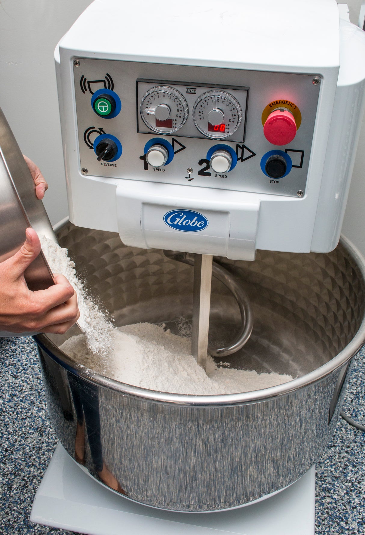 A person pours flour into a large spiral dough mixer with a 130 lbs capacity. The industrial mixer features a stainless steel bowl, visible mixing arm, control panel with dials and buttons, red emergency stop, and the “Globe” brand on the front.