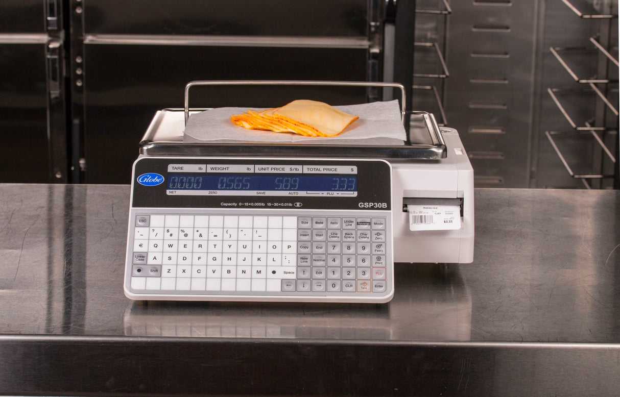 A legal for trade digital deli scale displays 0.565 pounds with sliced cheese on parchment atop its tray. Its keypad, dual-sided LCD display, and label printing scale feature are set against stainless steel surfaces and racks.