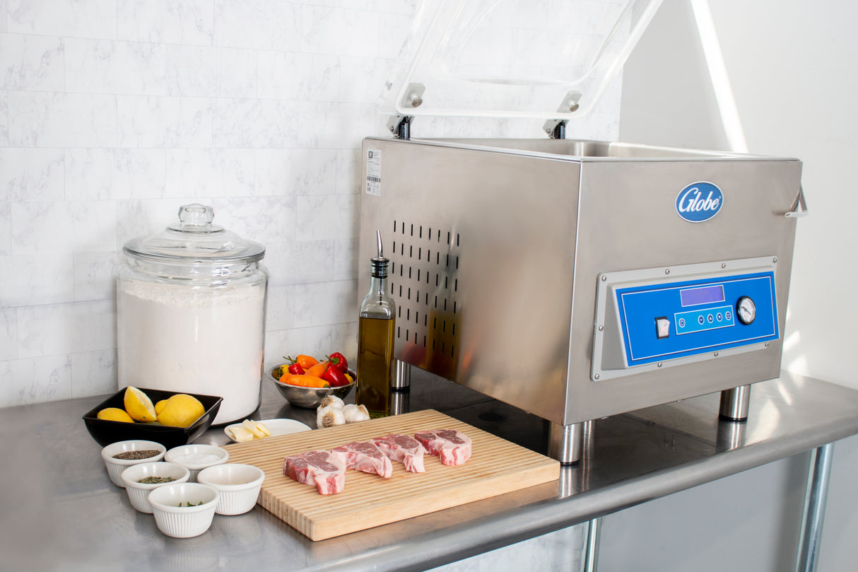 A stainless steel kitchen counter displays a Vacuum Packaging Machine, raw lamb chops on a wooden cutting board ready for marination programming, small bowls of spices, garlic, olive oil, lemons, bell peppers, and a large jar of flour against a white tile wall.