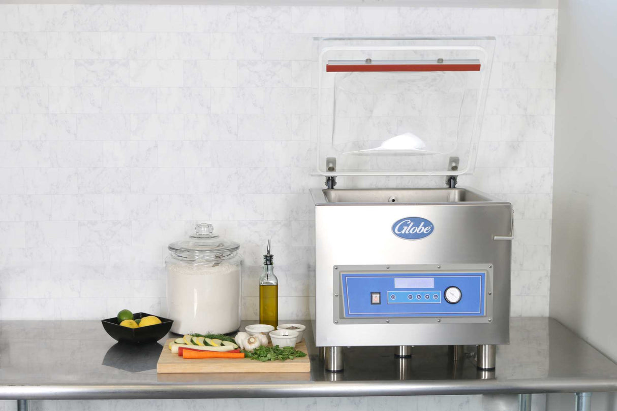 A stainless steel Vacuum Packaging Machine with the lid open sits on a metal counter. Surrounded by a jar of flour, citrus fruit, olive oil, and fresh produce, it features marination programming. The background is a light marble-patterned wall.