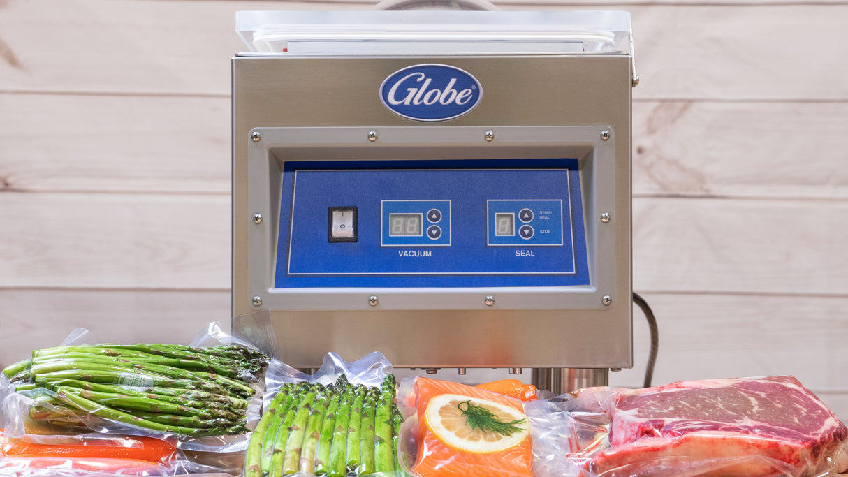 A stainless steel Globe GVP6 vacuum packaging machine with a blue control panel and 10-1/5" seal bar sits on a wooden surface. In front, vacuum-sealed bags hold asparagus, salmon with lemon, and raw steak. Light wood paneling forms the background.