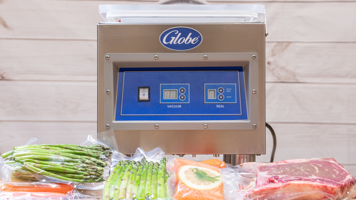 A stainless steel Globe GVP6 Vacuum Packaging Machine with a 10-1/5" seal bar and blue control panel is shown. In front, vacuum-sealed bags contain asparagus, a salmon fillet with lemon slice, and a raw steak. Light wooden panels form the background.