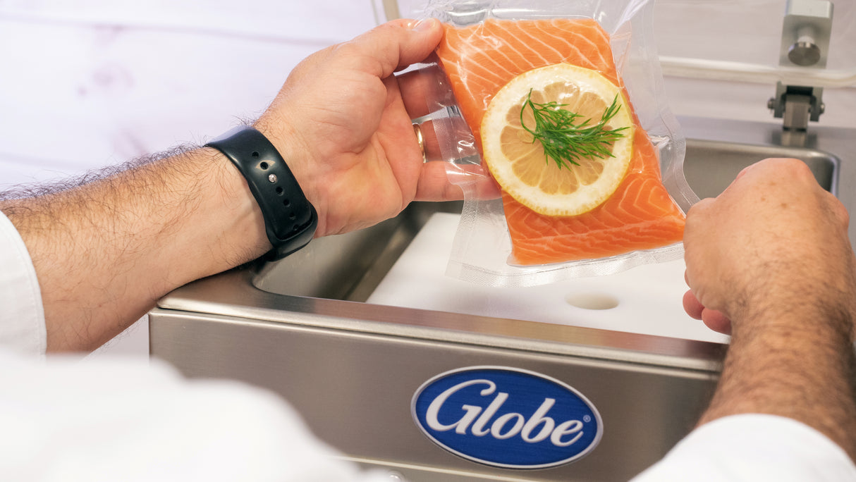 A person in a black wristband holds a vacuum-sealed salmon fillet with lemon and dill beside a stainless steel Globe GVP6 Vacuum Packaging Machine, featuring a 10-1/5" seal bar. The background is light and softly blurred.