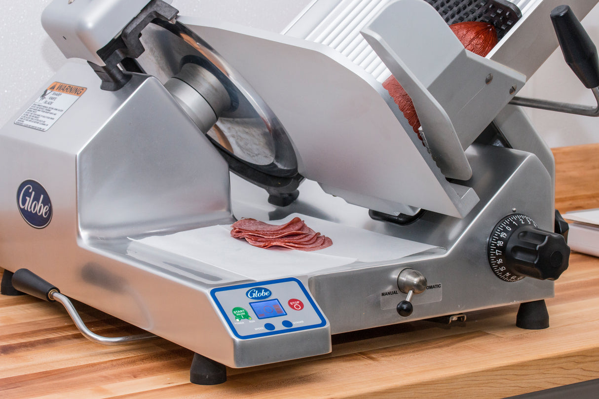 A premium heavy duty slicer sits on a wooden counter with thin slices of meat on its tray. The metallic automatic slicer features a sharp 13 inch steel knife, adjustable thickness dial, and digital controls, with part of a chunk of meat on the carriage.