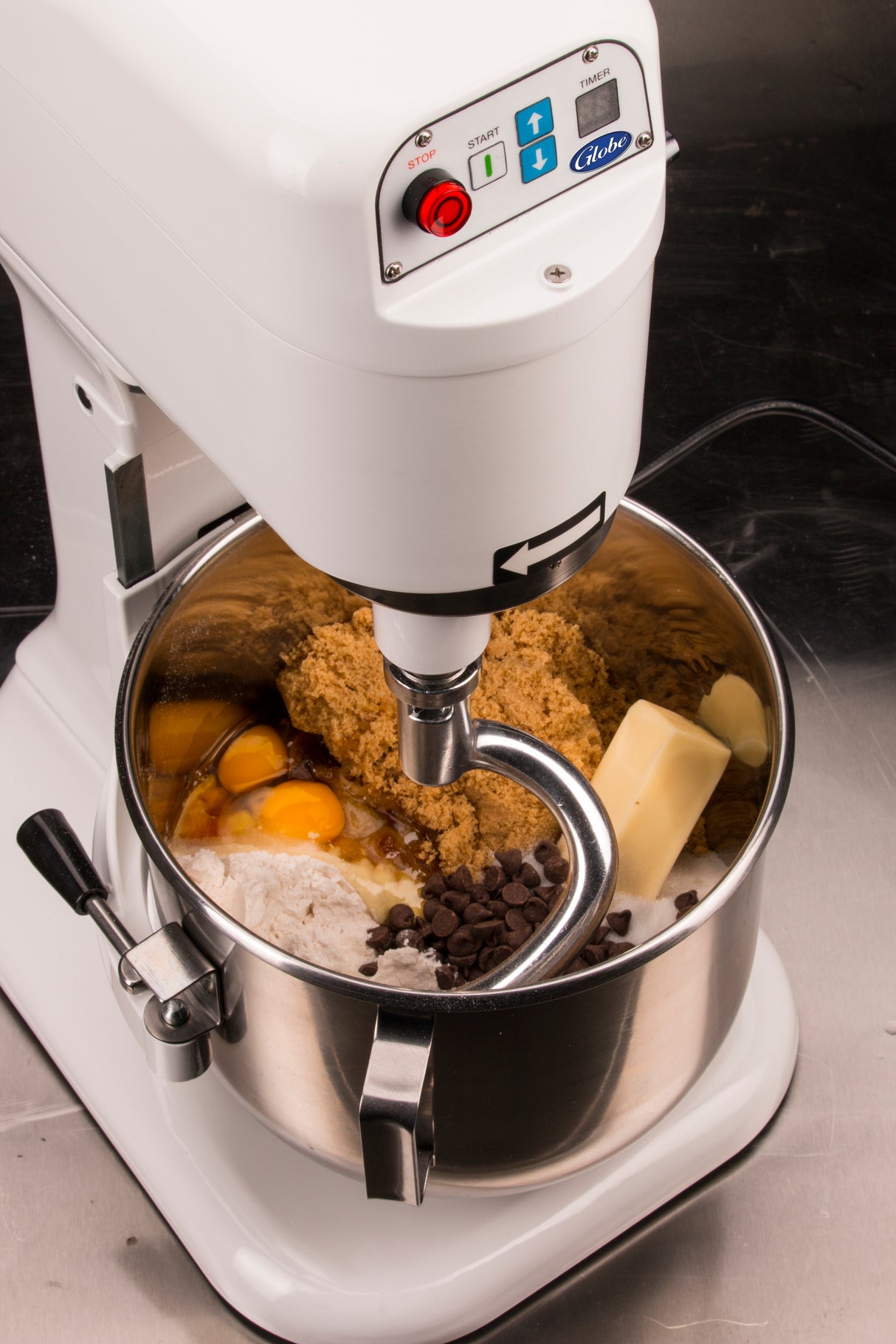 A white countertop mixer with a dough hook attachment holds eggs, brown sugar, flour, butter, and chocolate chips for cookies. The planetary mixer sits on a dark counter, ready to blend the ingredients to perfection.