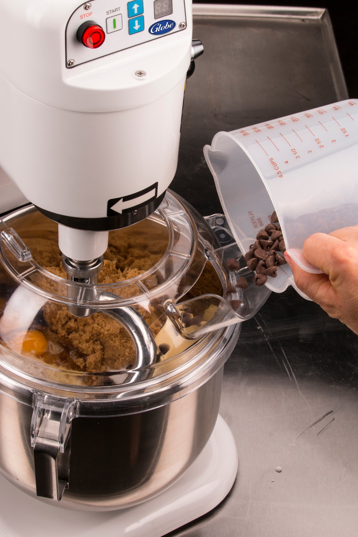 A person pours chocolate chips from a plastic measuring cup into a countertop mixer bowl with brown sugar and eggs. The 8 qt mixer, on a stainless steel counter, is running with its lid partially open to add the ingredients.