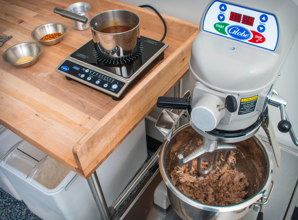 A 20 qt mixer with shredded meat inside sits on a counter next to a wooden prep table holding a hot plate with a pot of brown liquid and small metal bowls containing spices, suggesting food preparation in a commercial kitchen.