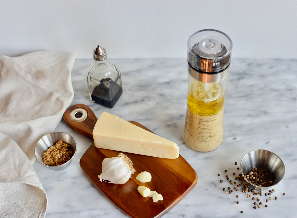A wedge of cheese, whole garlic bulb with peeled cloves, jar of oil, vinegar bottle, and grainy mustard in a 6 oz stainless steel sauce cup sit on a marble surface with a beige cloth, ready for meal prep.
