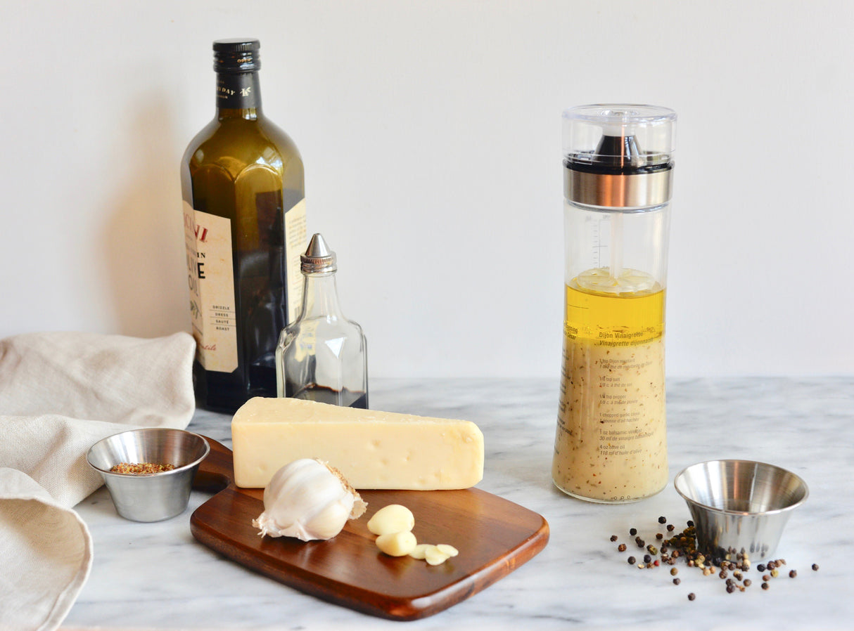 A marble countertop with a bottle of olive oil, a small glass vinegar bottle, a block of cheese, garlic cloves, a dressing shaker filled with vinaigrette, two 6 oz stainless steel sauce cups with spices, and scattered black peppercorns. A beige napkin sits on the left.