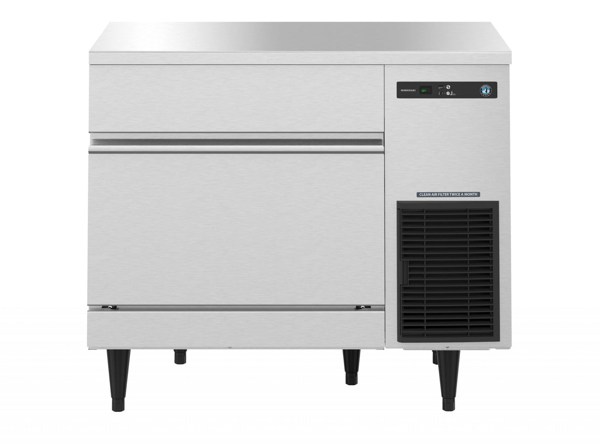 A Hoshizaki undercounter ice maker with a flat top, digital control panel, and lower right ventilation grille. This cube-style ice maker stands on black legs, features a left-side ice retrieval door, and boasts a clean, reflective stainless steel surface.