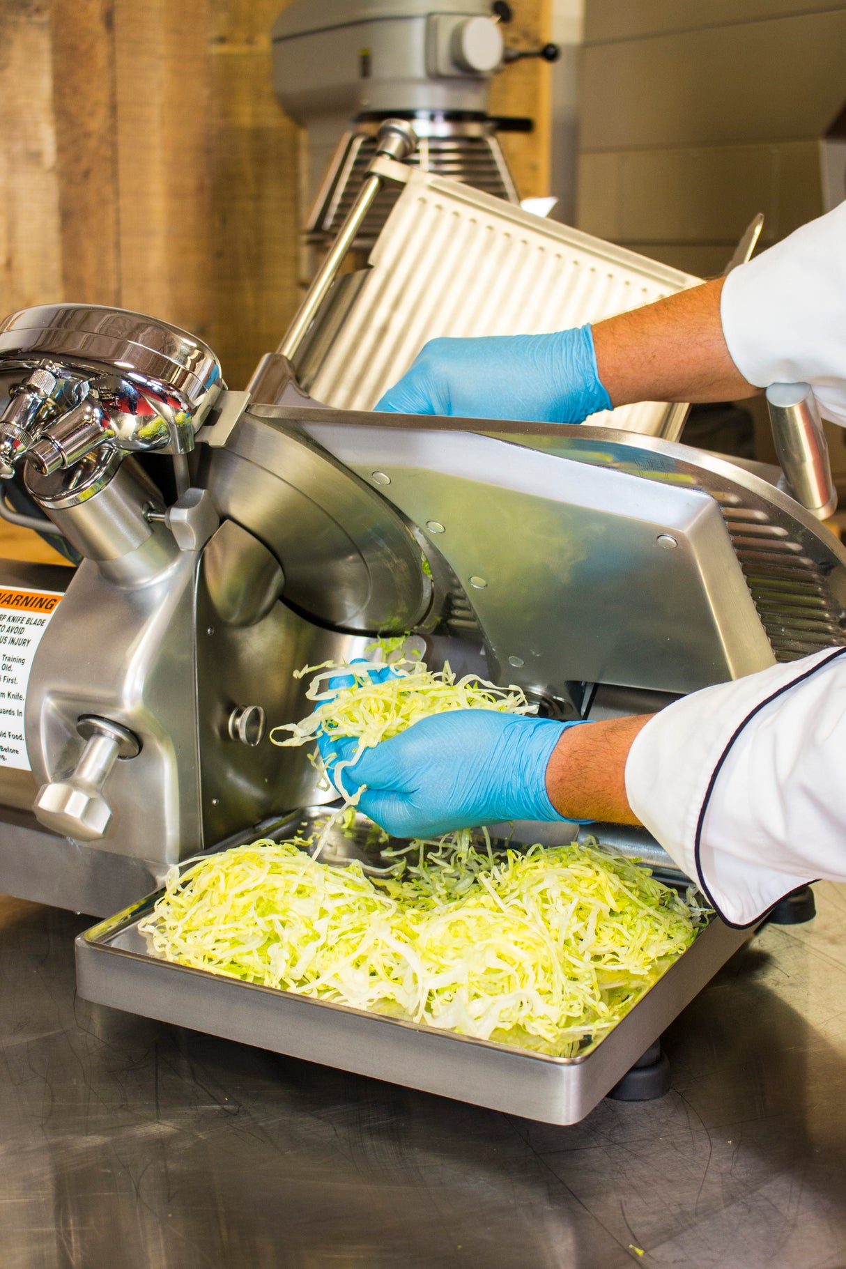 A person wearing blue gloves and a white chef’s coat uses a Chefmate Series compact slicer to shred cabbage, which falls into a tray below, in a commercial kitchen setting.