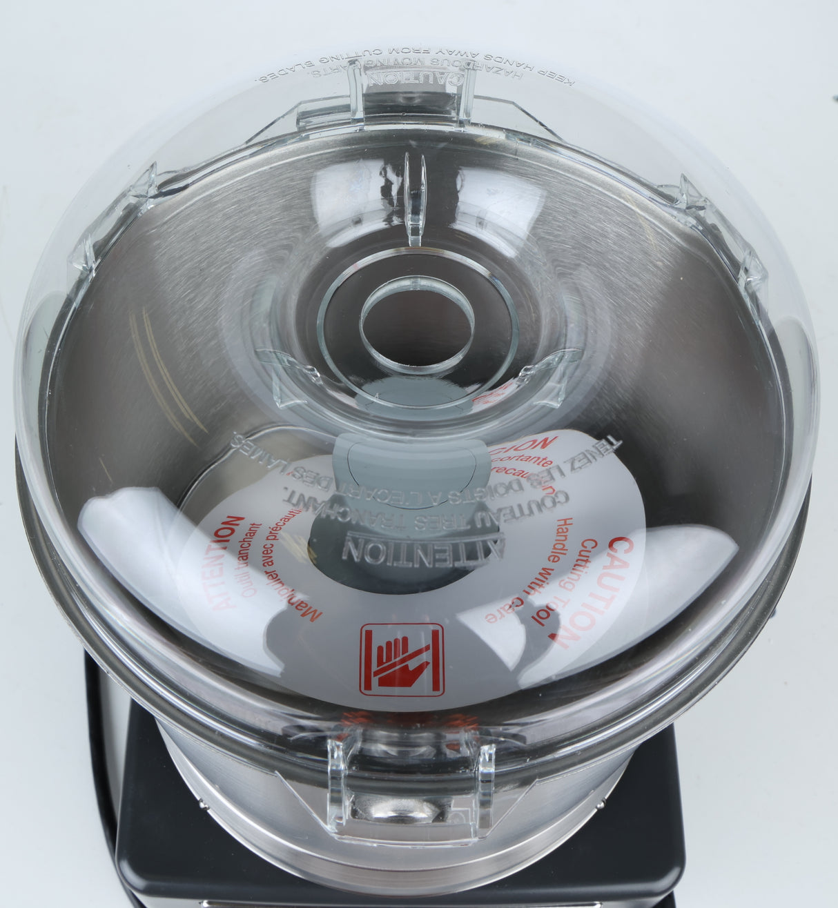 Overhead view of a Robot Coupe food processor with a transparent lid. Inside is a metal bowl and a sharp S-shaped stainless steel blade. Red caution labels warn against contact with the blade. The commercial food processor sits on a black base against a plain white background.