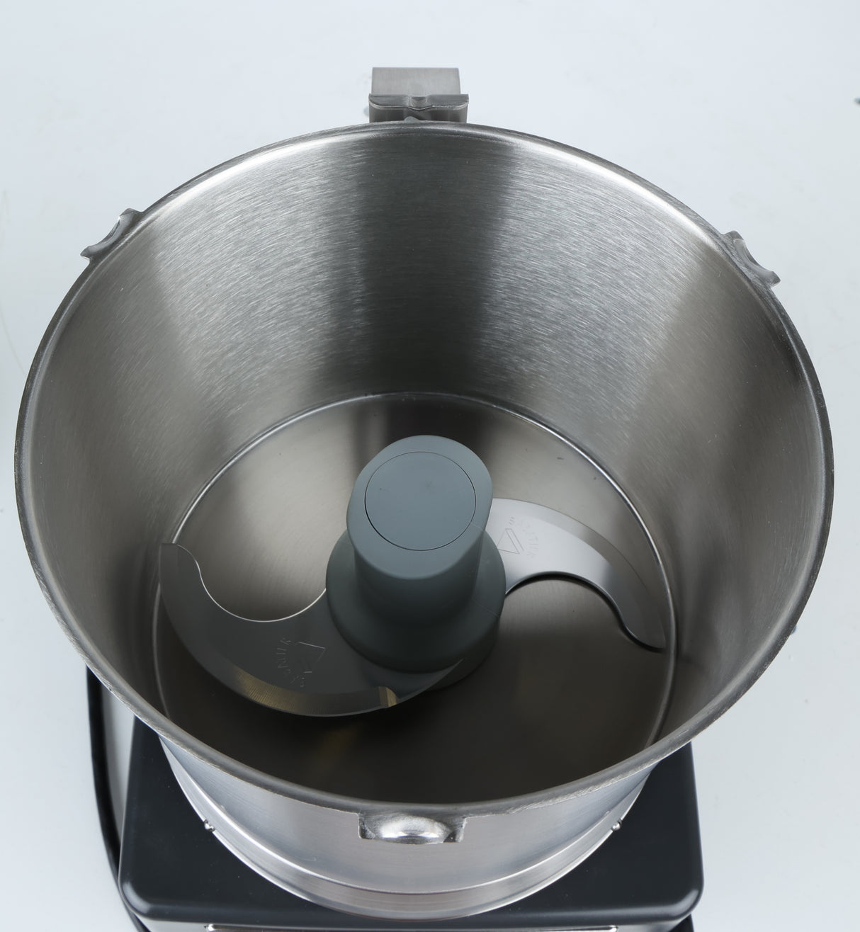 A top-down view of an empty stainless steel commercial food processor bowl with a sharp metal S-blade attachment in the center, set on a black base against a white background. The bowl, compatible with a Robot Coupe food processor, features a brushed metal texture and two side handles.