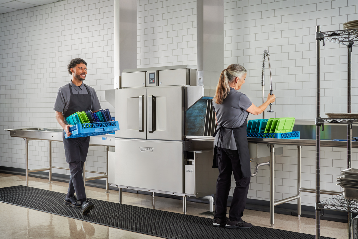 Two people in gray uniforms work in a commercial kitchen: one carries a blue rack of trays, smiling, while the other rinses trays before loading them into a conveyor dishwasher. The background is white tiled walls and shelves with clean plates.