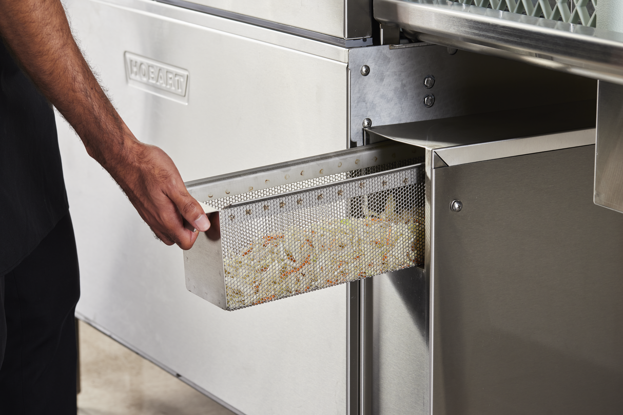 A person’s hand pulls out a perforated metal basket filled with shredded vegetables from a stainless steel high capacity dishwasher. The appliance has the brand name “Hobart” visible on its front.