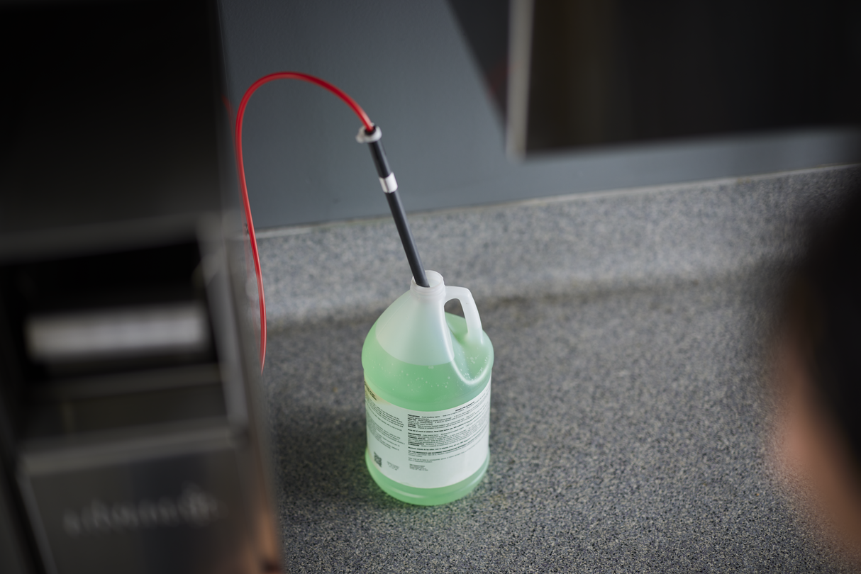 A large, translucent green plastic jug of cleaning solution with a white handle sits on a gray countertop. A black and red tube is inserted into the cap, suggesting use with a Hobart CL66VL-4 ventless energy recovery conveyor dishwasher. The background is out of focus.