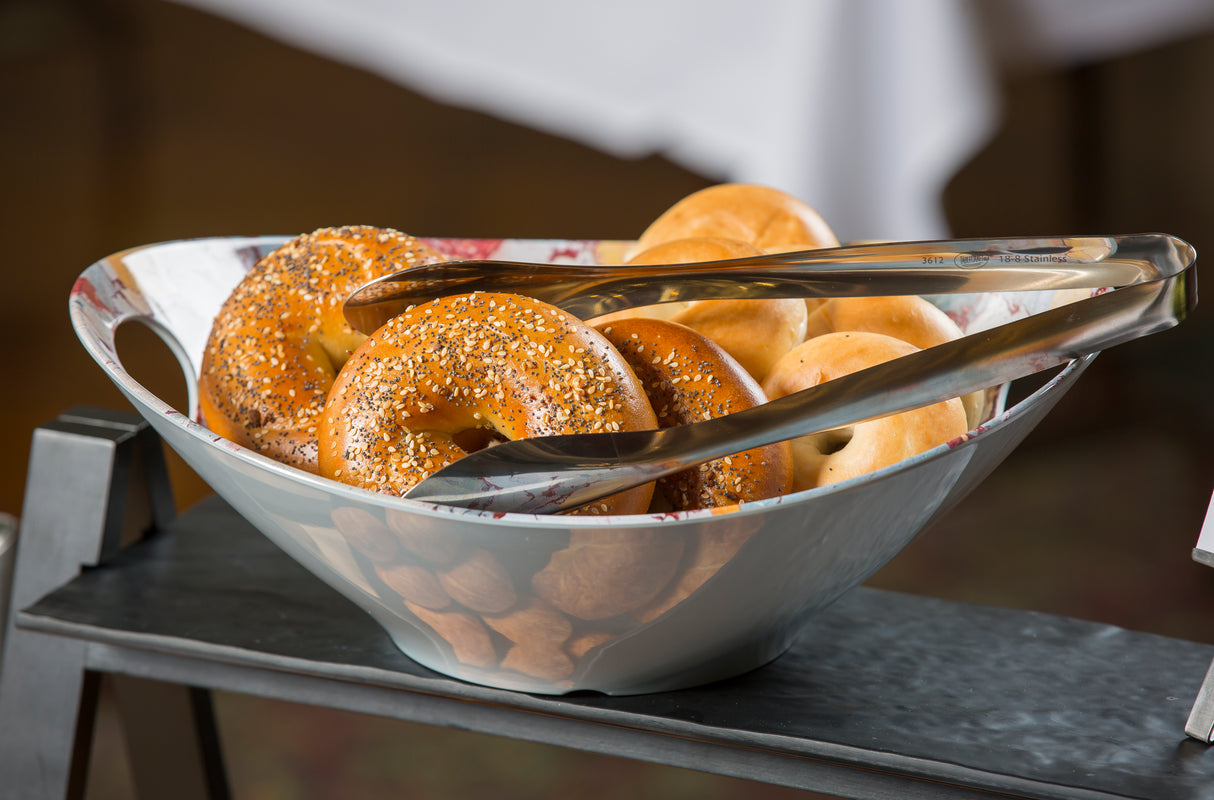 A bowl from the Dalton II Collection holds assorted bagels, with a stainless steel tong server resting on top. The bowl sits on a dark tray, against a blurred background and white tablecloth. All items are dishwasher safe for easy cleanup.