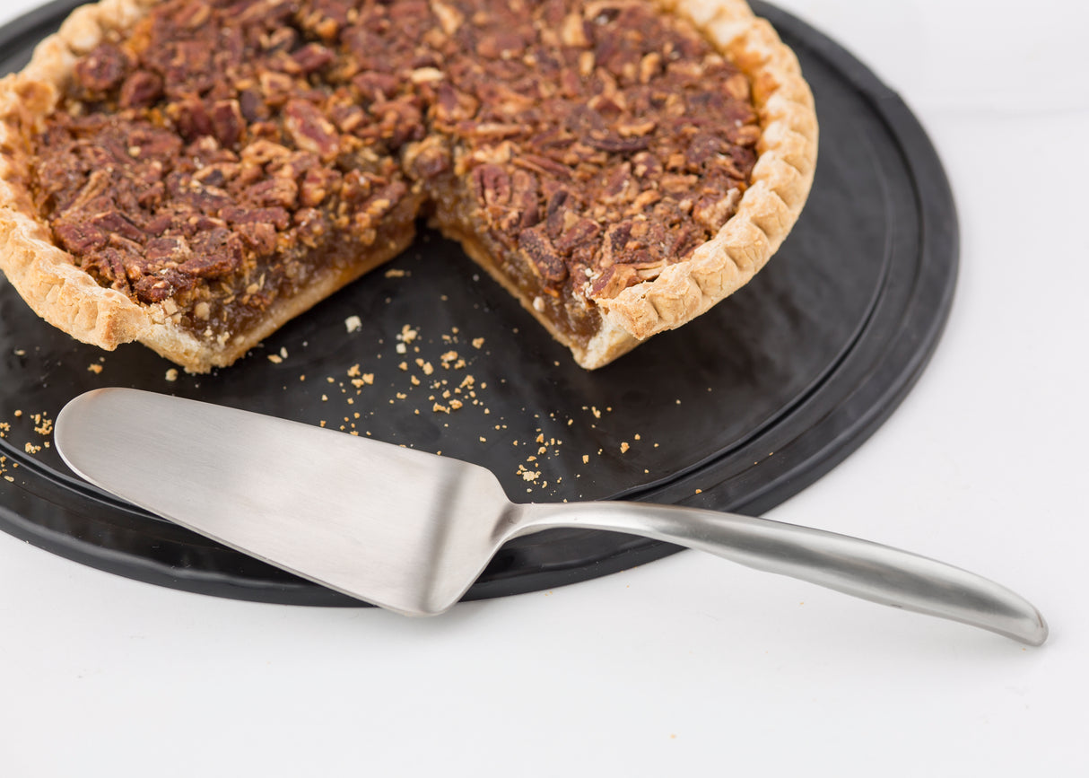 A pecan pie with a slice missing sits on a black tray. An 18/8 stainless steel Buffet Cake Server from the Dalton Collection rests in front of the pie, which has a golden crust and whole pecans atop its rich filling. Crumbs are scattered on the tray.