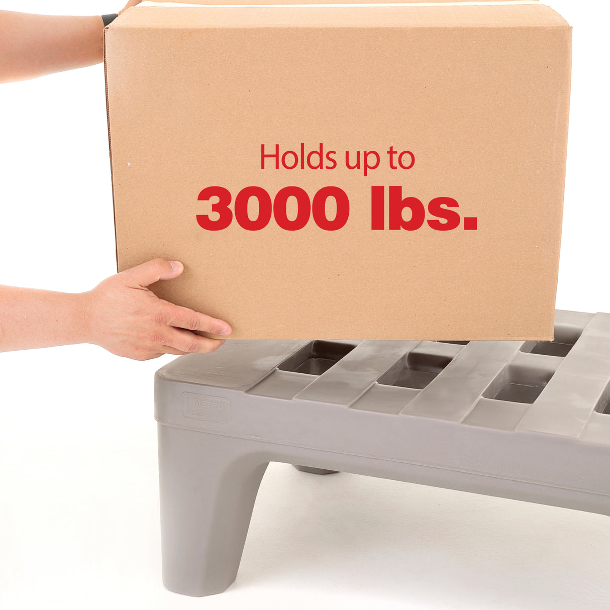 A person places a large cardboard box labeled “Holds up to 3000 lbs.” on a corrosion proof dunnage rack with four sturdy legs and slotted openings, against a plain white background.