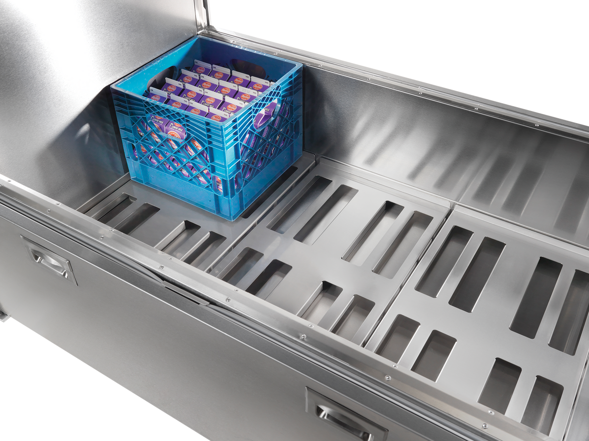 A blue plastic crate filled with individual cartons of milk sits inside a large, empty stainless steel milk cooler with slotted metal shelving and forced-air circulation. The crate is positioned near one end of the unit.
