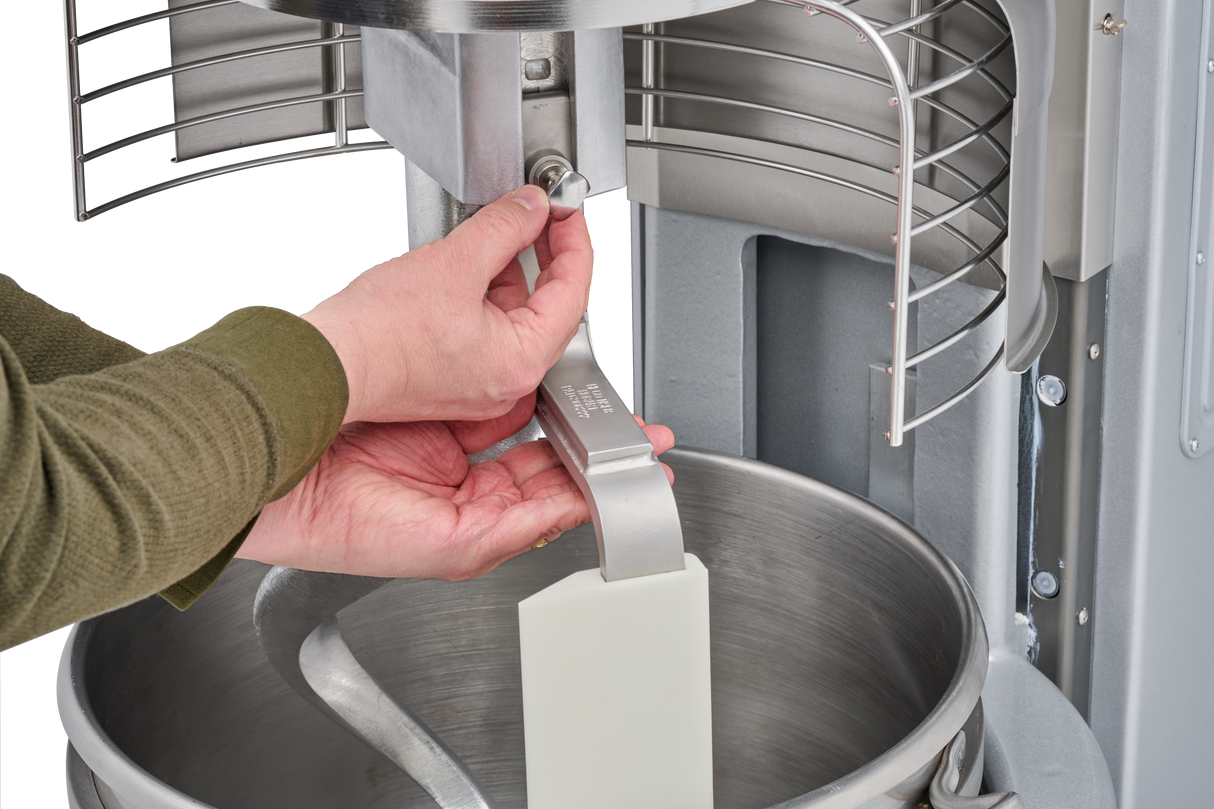 A person’s hands attach a metal tool inside a 60 quart mixer, locking it in place above a large metal mixing bowl with a spiral dough hook and safety guard visible. The person wears a green long-sleeve shirt.