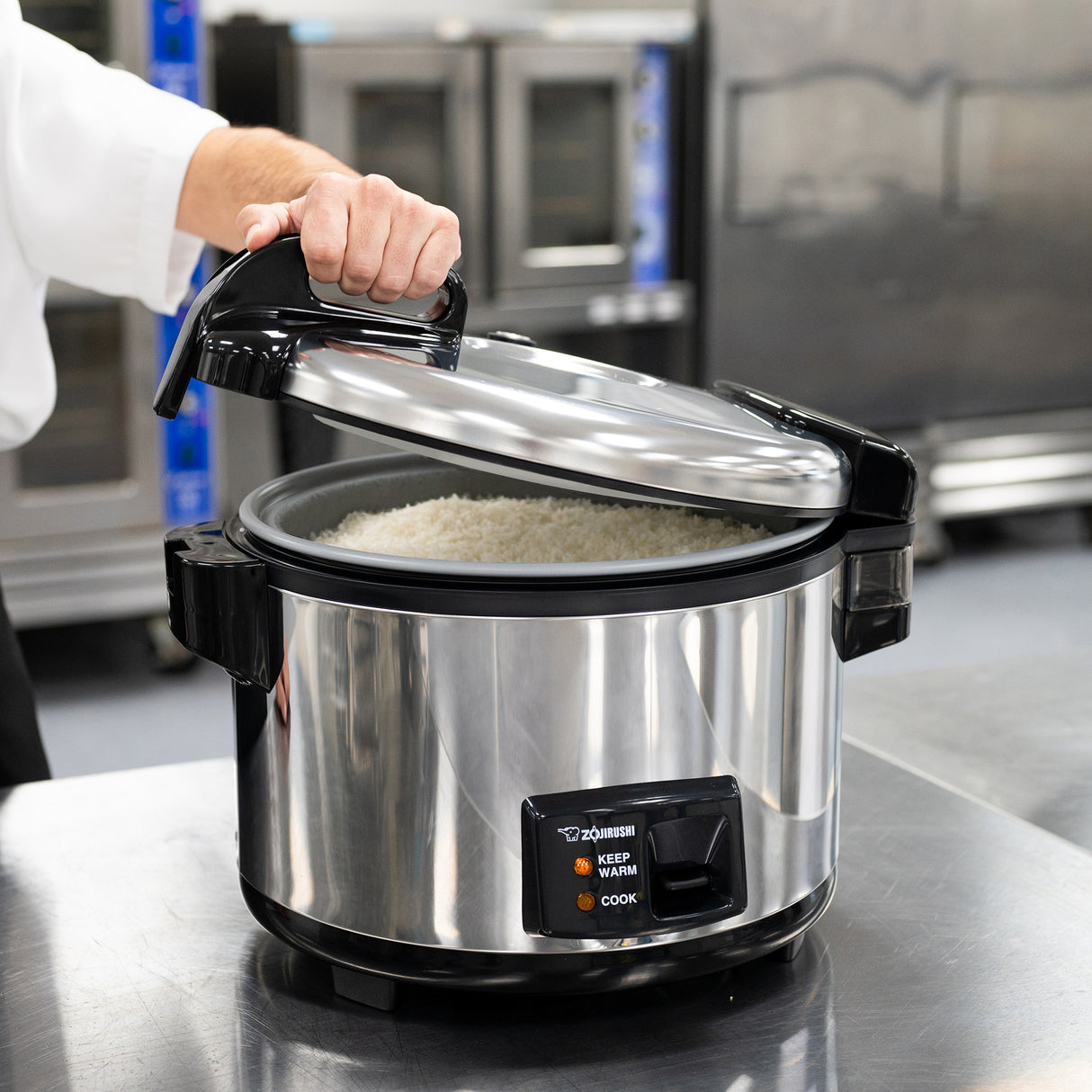 A person in a white sleeve lifts the lid of a Zojirushi Commercial Rice Cooker filled with cooked white rice. The 20 cup capacity cooker sits on a metal countertop, its control panel showing “Keep Warm” and “Cook” buttons.