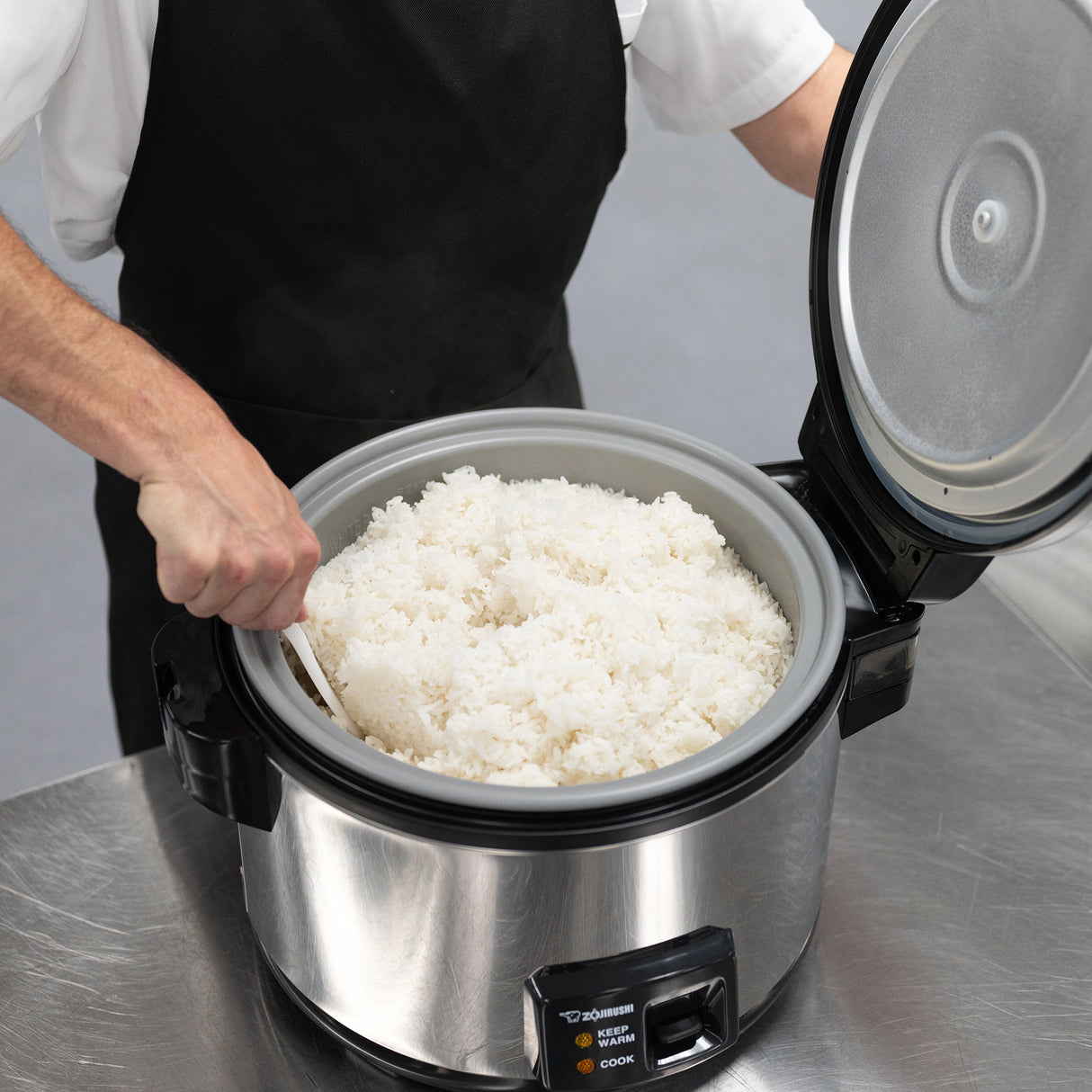A person in a black apron and white shirt fluffs white rice with a plastic paddle inside a stainless steel commercial rice cooker, featuring a 20 cup capacity, on a shiny metal countertop. The open lid reveals the inner pot full of fluffy rice.