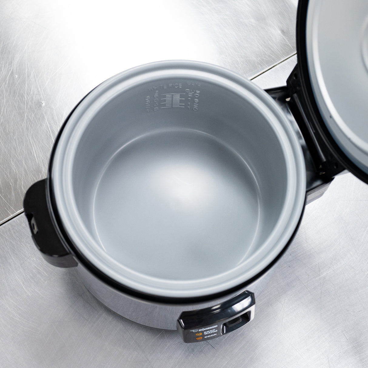 A top-down view of a commercial rice cooker on a stainless steel countertop. The stainless steel rice cooker’s inner pot is empty, with measurement markings visible inside. The digital control panel and partially open lid are also shown.