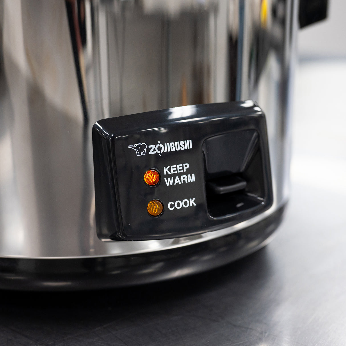 Close-up of a Zojirushi stainless steel rice cooker control panel with two indicator lights—KEEP WARM (orange) and COOK (red). The black panel features the brand logo, and the body hints at its commercial rice cooker 20 cup capacity.