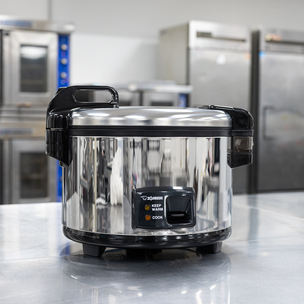 A shiny Zojirushi Commercial Rice Cooker with a 20 cup capacity and black handles sits on a reflective metal countertop in a commercial kitchen, its control panel labeled “Keep Warm” and “Cook,” surrounded by industrial appliances.
