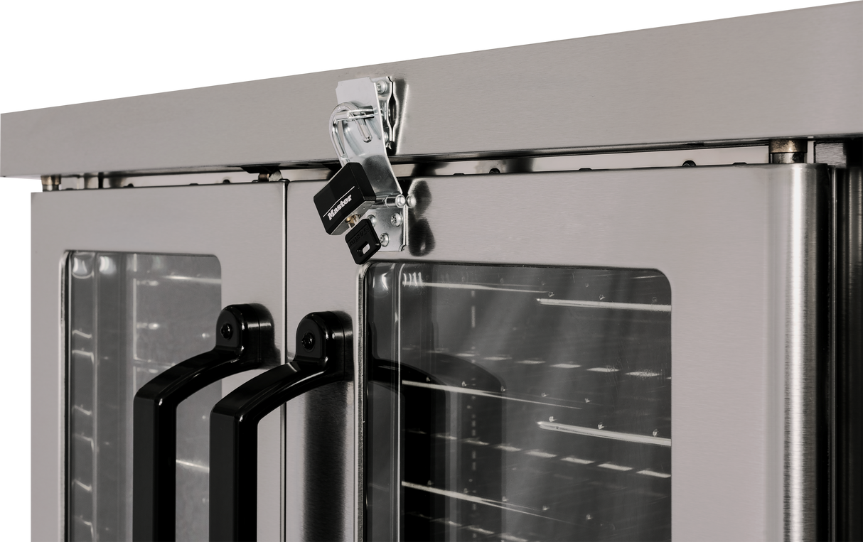 Close-up of a stainless steel double-door commercial refrigerator with black handles and glass panels, positioned beside a gas double-deck convection oven. A metal latch with a padlock secures the doors shut, revealing interior shelves through the glass.