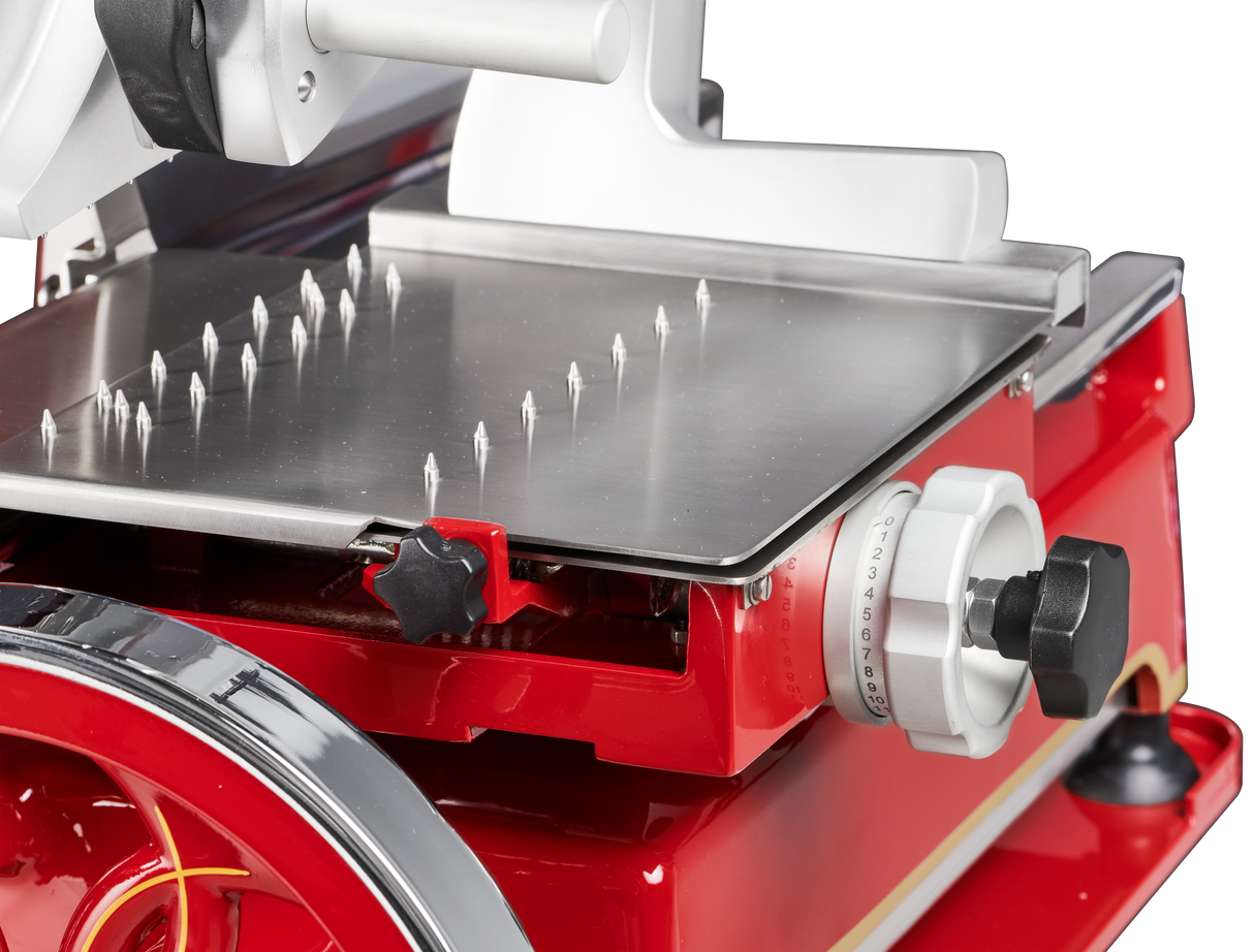 Close-up of a Berkel Fly Wheel Slicer shows a shiny metal platform with sharp spikes for holding food, a white adjustment dial with numbers, and various knobs. The chromium-plated carbon steel knife gleams against the black background.