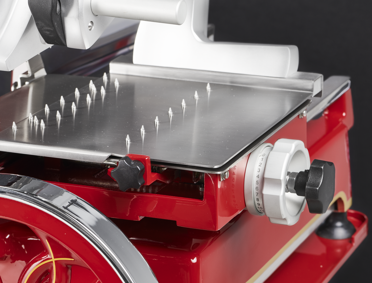 Close-up of a red and silver Berkel Fly Wheel Slicer, focusing on the stainless steel platform with pointed spikes, adjustment dial, and part of the chromium-plated carbon steel knife. The black background highlights the machine’s details and metallic textures.