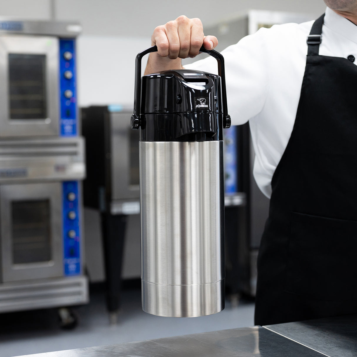 A person in a white shirt and black apron holds a large Zojirushi Airpot coffee dispenser with a stainless steel vacuum liner for superior heat retention, in a commercial kitchen with ovens and metal surfaces visible in the background.