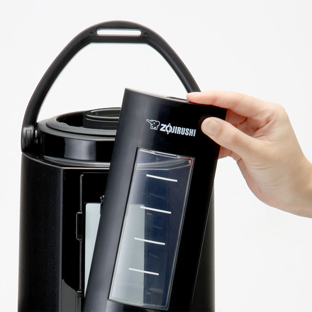 A hand is removing the black lid with a clear window and white measurement markings from an insulated Zojirushi beverage dispenser, featuring a stainless steel vacuum liner and handle, against a plain white background.