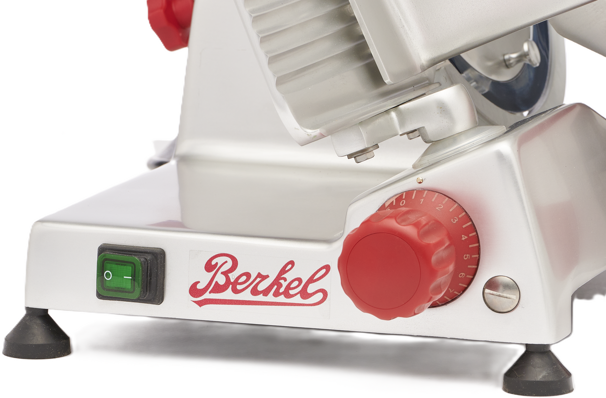 Close-up of a Berkel B10-SLC gravity feed slicer’s base, showing a green power switch, the red Berkel logo, a large red dial for thickness adjustment, metallic housing, and four black rubber feet for stability.