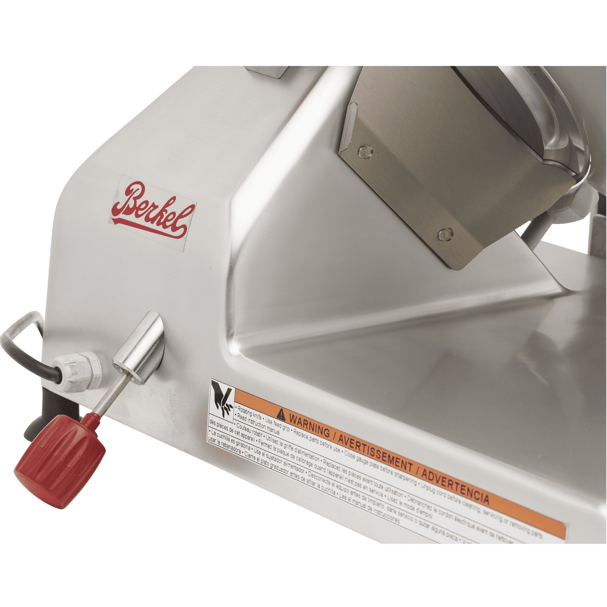 Close-up of a silver Berkel slicer, a manual gravity feed slicer, showing the brand logo, red-handled control knob, metal lever, and an orange warning label with text in English, French, and Spanish on the machine’s shiny metal surface.