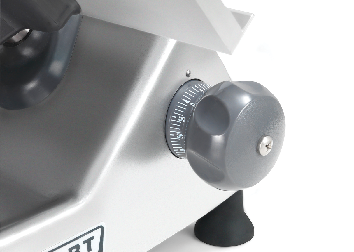A close-up of a gray adjustment knob with numbered markings on a metallic Heavy Duty Meat Slicer, likely a manual meat slicer. The knob is set near 15 and attached to an angled surface, with rubber feet visible underneath for stability.