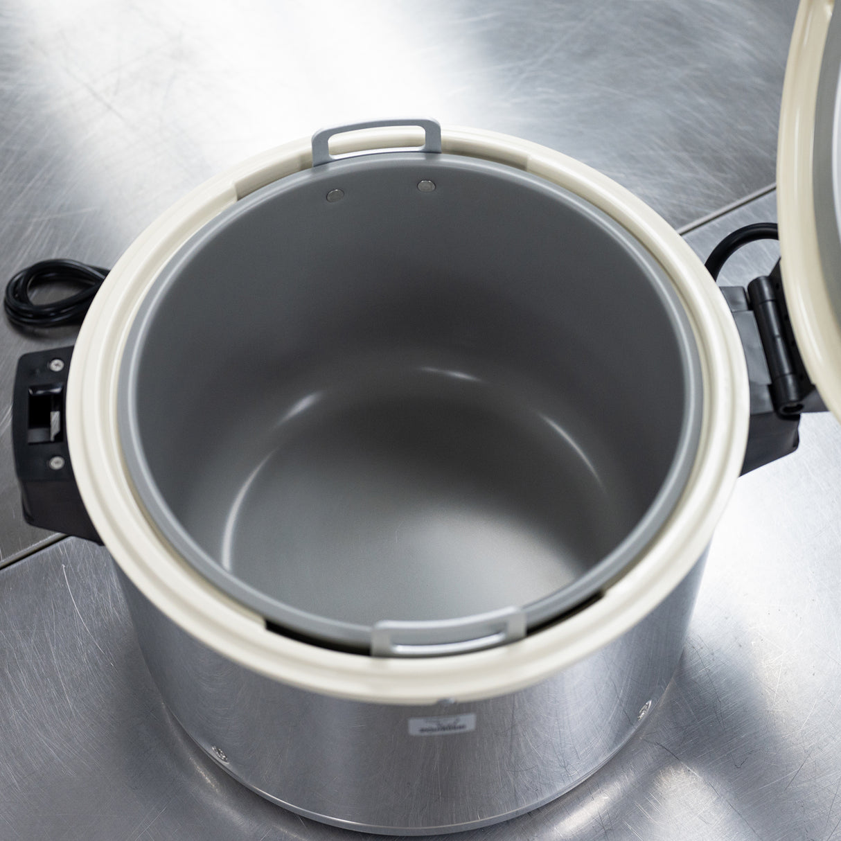A large, open commercial rice cooker sits on a reflective stainless steel surface. The cooker’s interior is clean and empty, featuring a gray nonstick inner pot. The white lid is open, and a black power cord is visible on the left side.