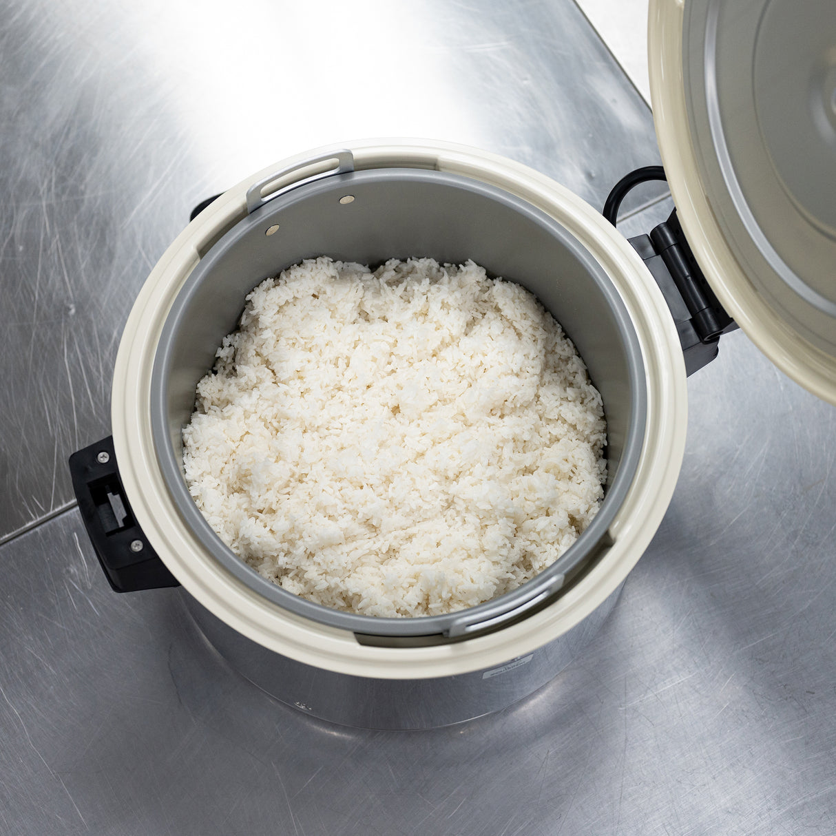 A Zojirushi America commercial rice cooker with its lid open sits on a metal countertop, revealing freshly cooked white rice inside. The gray interior pot holds fluffy, evenly cooked rice as light reflects off the shiny surface of the counter.