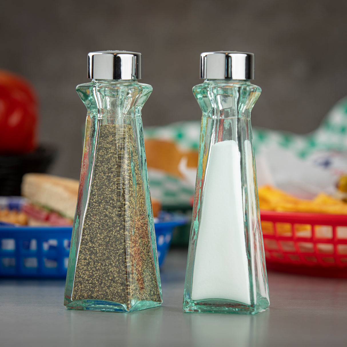 Two Marbella Shakers with chrome plated ABS tops are filled with black pepper (left) and white salt (right). Made from recycled green tint glass, they sit side by side on a table, with blurred food baskets and a red object in the background.