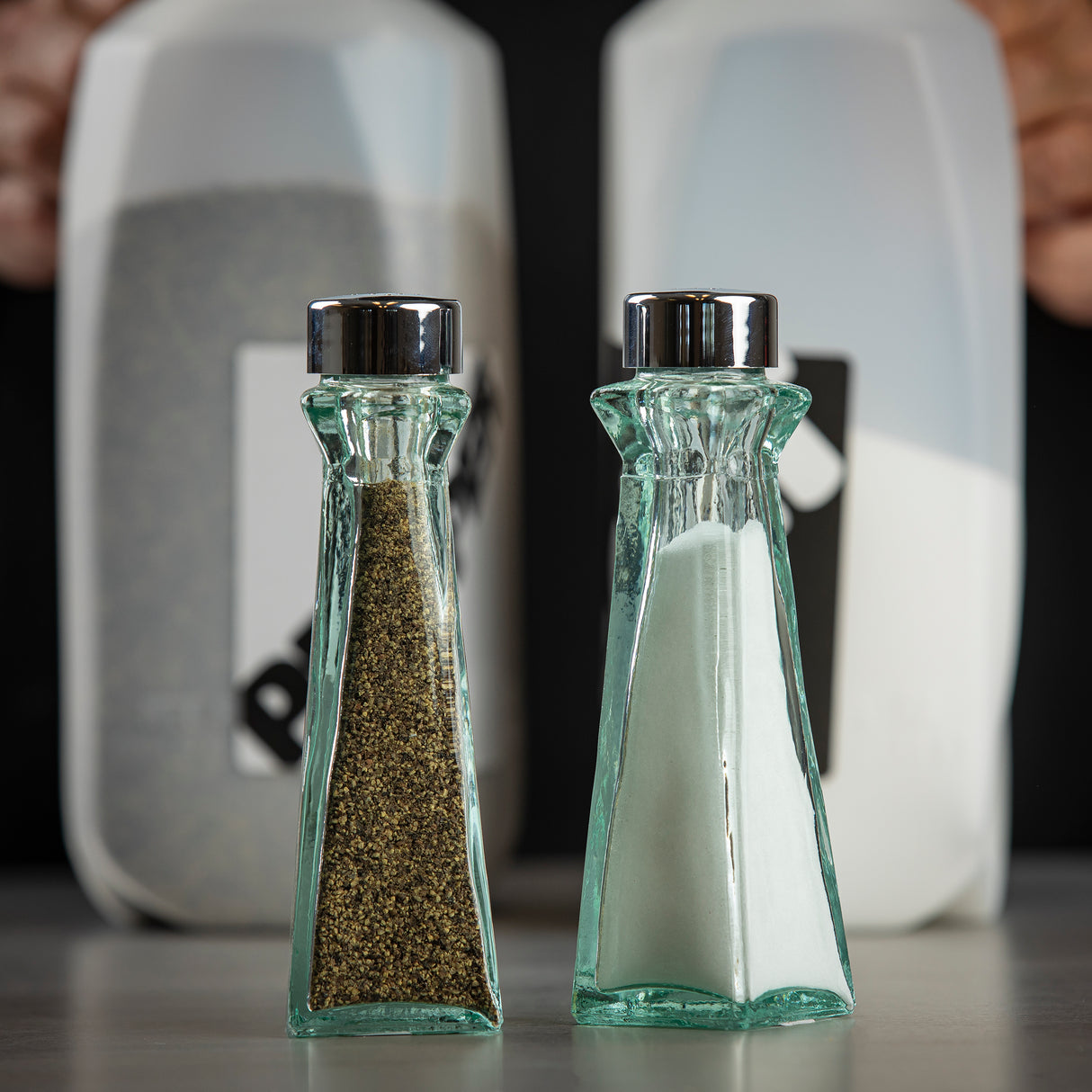 Two Marbella Shakers with chrome plated ABS tops, one filled with black pepper and one with white salt, stand side by side on a surface. The shakers feature recycled green tint glass, while large labeled containers sit blurred in the background.