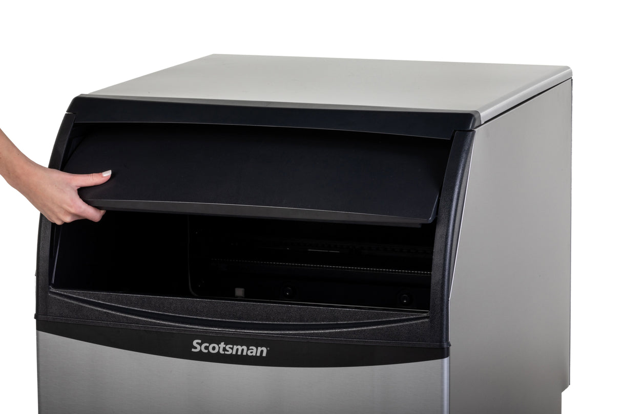 A person’s hand opens the black lid of a stainless steel Scotsman ice machine against a white background. The modern, industrial undercounter ice maker reveals its dark, empty compartment and sleek design.