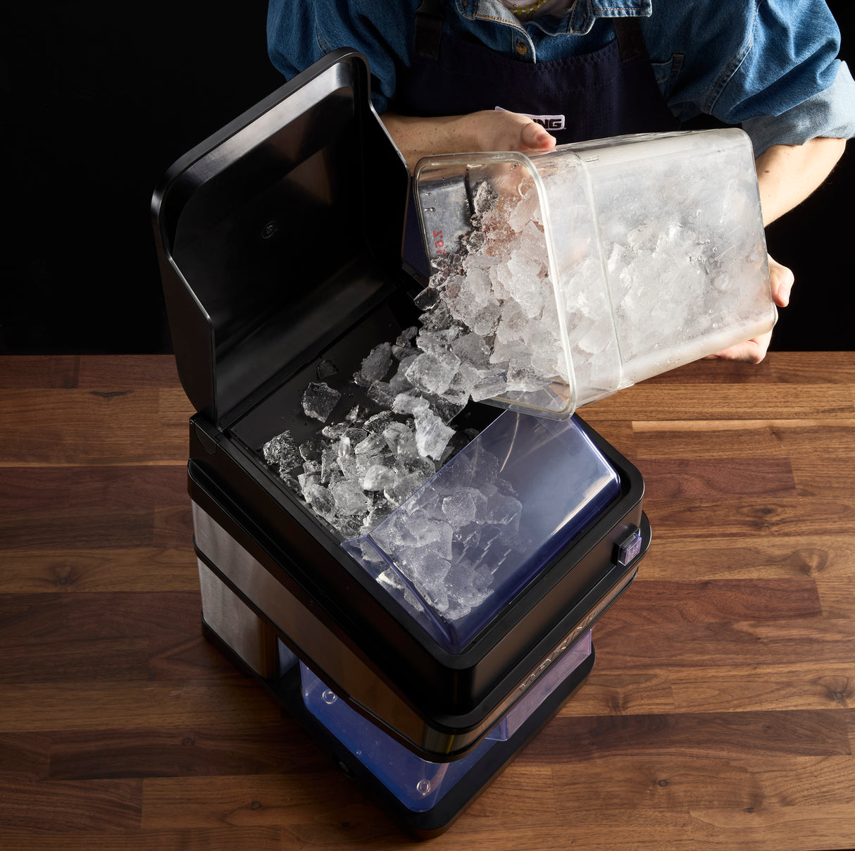 A person in a denim shirt pours a clear container of ice cubes into the open top of a black Waring Ice Crusher, which sits on a wooden surface with some ice already inside the machine.