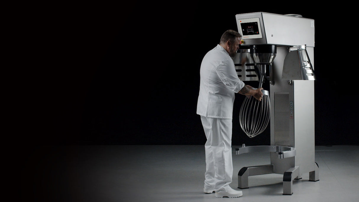 A man dressed in all white operates a heavy duty planetary mixer with a metal whisk attachment, set against a dark background. The stainless steel mixer features a digital control panel as he stands facing it, adjusting the whisk.