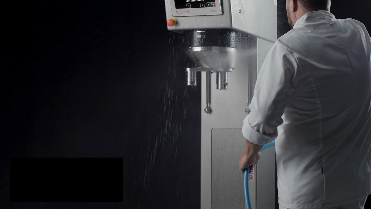 A person in a white chef’s coat sprays water onto a large industrial planetary mixer with a hose. The metallic machine, equipped with control buttons on top, gleams with water droplets contrasting against the dark background. The person’s face is not visible.