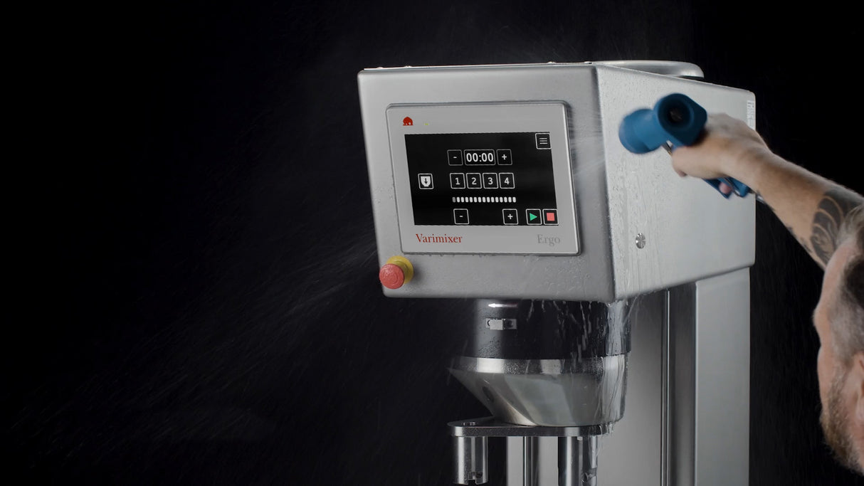 A person sprays water on the control panel of a Varimixer Ergo heavy duty planetary mixer against a black background, demonstrating that the programmable mixer is resistant to water and can be safely washed down.