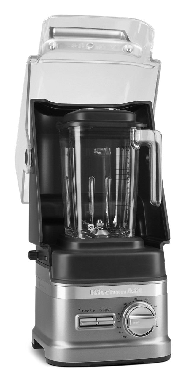A silver KitchenAid Commercial Blender with a 60 oz. capacity sits inside a transparent sound enclosure, lid open. The clear pitcher and handle are visible, and the control panel features a dial and buttons on a white background.