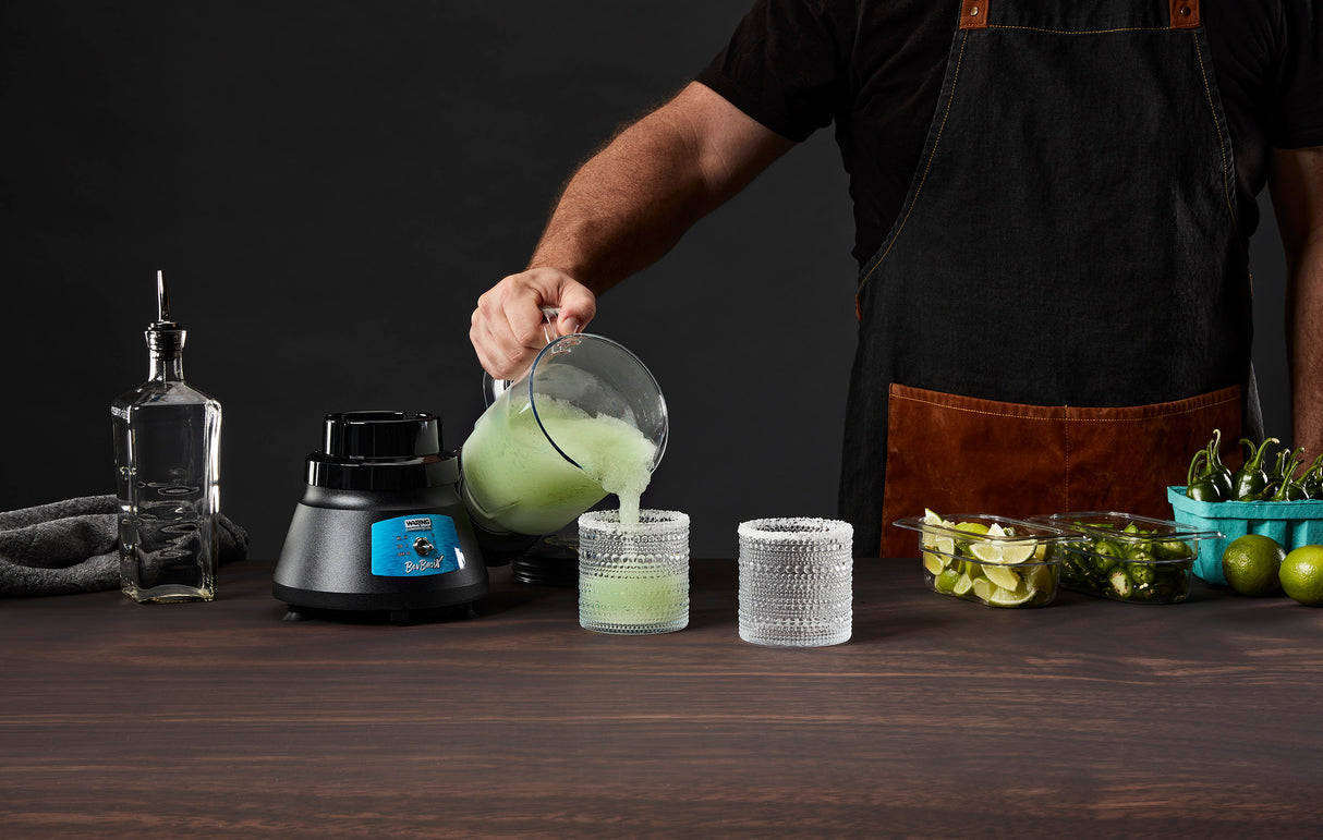 A person in a black apron pours a green smoothie from a blender with a stainless steel blade into two textured glasses on a wooden table. Nearby are the Bar Blender base, bottle, towel, limes, sliced limes, and green chili peppers against a dark background.